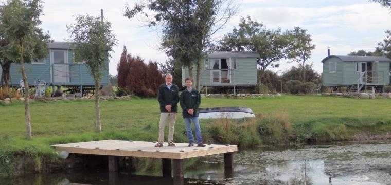 Mark Barrowman and Jonathan Gooders stood in front of their shepherd huts on the pontoon of a lake.