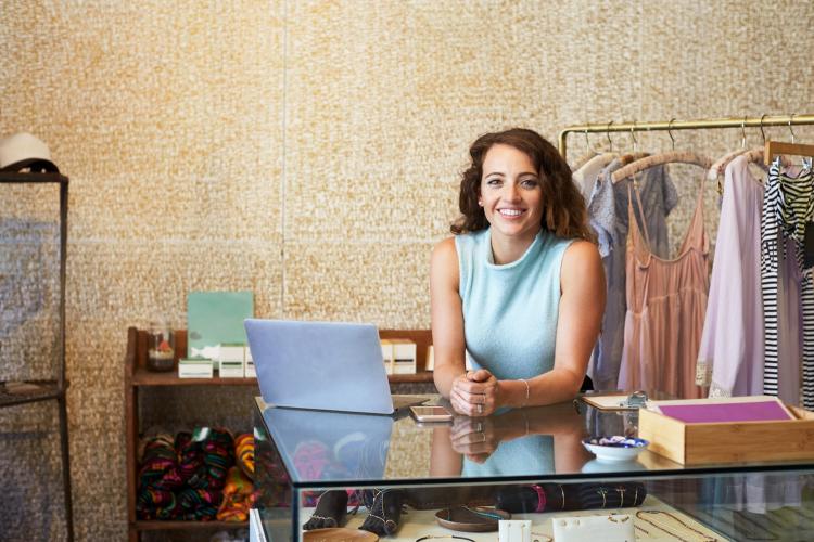 young female business owner working in clothes shop, smiling for photo and leaning on desk 